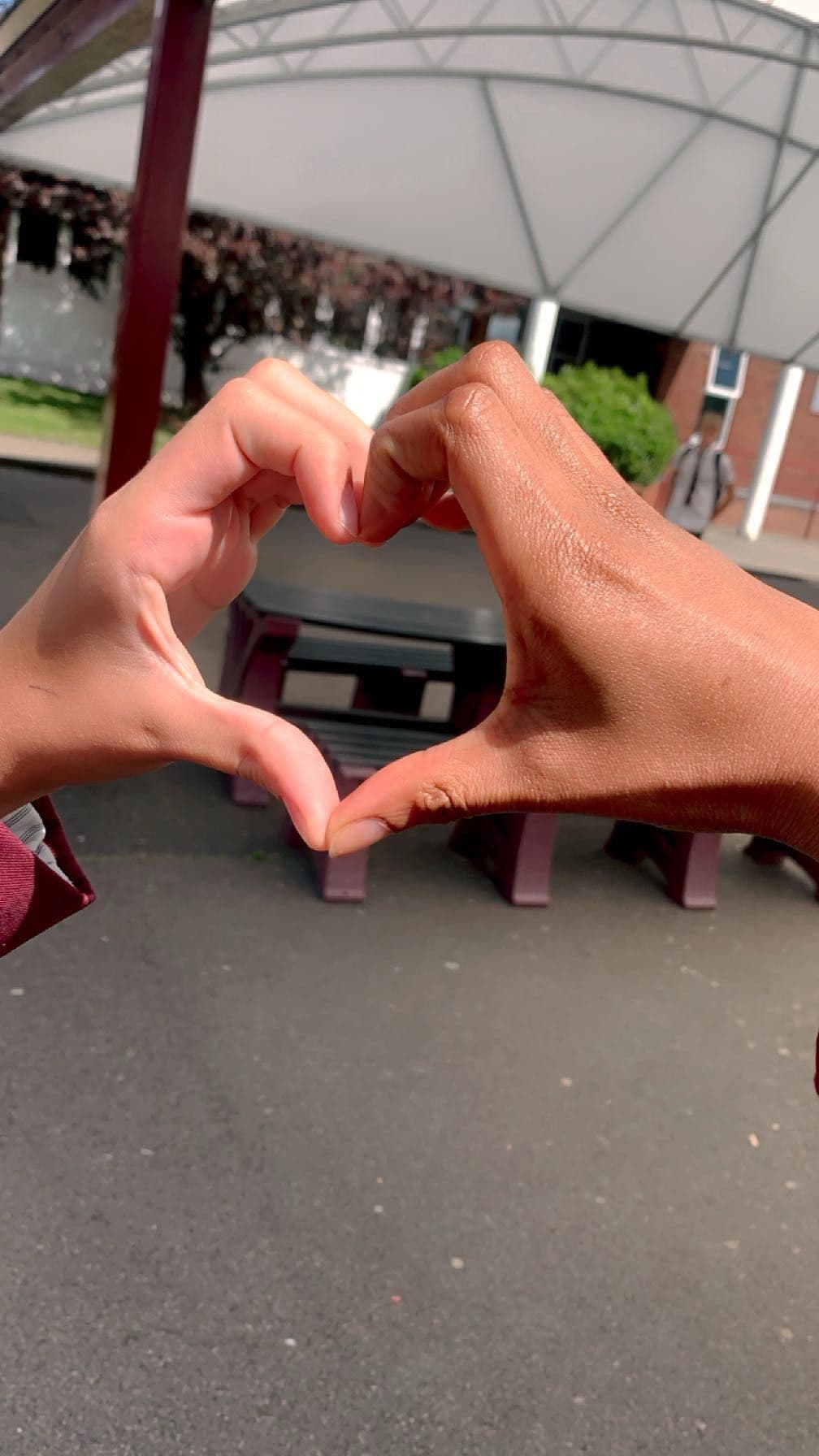 Two hands of different skin tones join together to form a heart shape outdoors.
